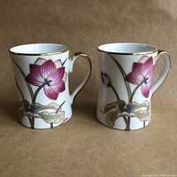 Pair of white porcelain mugs with pink and gold cloisonne lotus floral design, gold rims and handles, standing side by side on a brown surface.