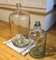 Large clear glass carboy and smaller blue-green tinted glass carboy with metal handle, shown side by side on a cloth on wooden floor