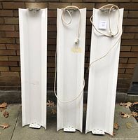 Three white metal grow-light fixtures lined up vertically against a brick wall. Each fixture is long and rectangular with shallow depth, containing bulb sockets at each end and visible power cords.