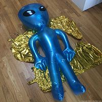 Blue inflatable alien figure laid on top of two uninflated gold mylar balloons on a wooden floor