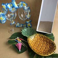 Overview of lot showing pineapple dish, stemmed glasses with lei, cocktail umbrellas, foliage, and box of sand with shells.