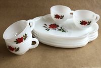 Photo showing the full set of four white milk glass oval plates stacked with four matching cups with red rose decals, arranged on a brown background.