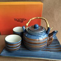 Set showing teapot with bamboo handle, two handle-less striped cups, rectangular striped tray, and Teaopia box in background.