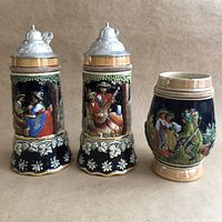 Photo showing front view of two lidded ceramic beer steins and one unlidded potbelly stein with hand-painted German folk scenes.