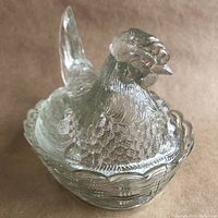 Close-up of the glass hen-shaped covered dish with detailed feathers, tilted head and split tail, sitting on a woven nest shape base.