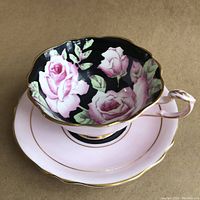 Cup nestled on saucer showing black interior with pink roses and blush pink exterior edged in gold