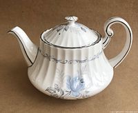 Side view of the white fluted teapot showing pale blue rose pattern with grey leaves and platinum trim, curved handle and spout, and domed lid with knob.