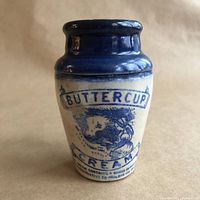 Front view of vintage stoneware cream pot with cobalt blue top and transfer print of milkmaid and cow labeled 'Buttercup Cream'.