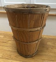 Front view of vintage tall bushel basket showing wood slats, metal bands, and metal hooks at top rim. Visible wear and some broken pieces on bottom section.