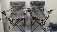 Two gray folding camping chairs set up side by side on a gray surface against a white wall.