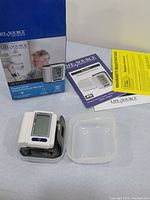 Digital wrist blood pressure monitor with wrist cuff, clear plastic storage case, manuals and warranty cards, all displayed on white surface along with original packaging box.