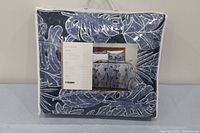 Front view of the coverlet set in packaging showing blue and white coverlet with leaf motif