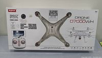 Front box view showing the Syma drone model D7000WH, features like 360-degree stunts and WiFi live streaming, recommended age 14+.