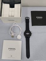 Fossil Gen 6 smartwatch with charger, manuals, and original box on light gray surface.