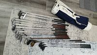 Overview of all golf clubs lined out on the floor beside the white and navy blue golf bag.