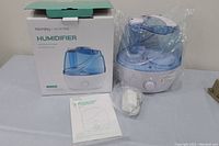Photo of a new Homasiy humidifier, still wrapped in plastic, with the product box, power adapter, and instruction manual beside it.