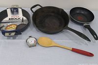 Photo of kitchen items including Starfrit Rock baking pan with packaging, cast iron skillet, non-stick frying pan, wooden spoon, and metal cooking thermometer.