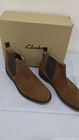 Pair of brown suede Chelsea boots with elastic side panels, displayed in front of the Clarks branded shoebox.
