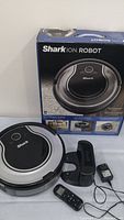Shark ION Robot floor cleaner displaying top view, box in background, charging dock, remote control, and power adapter visible.