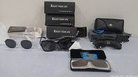 Photo showing six pairs of sunglasses arranged on a table with three black boxes labeled 'Enjoy Your Life', assorted cases including a Ray-Ban case, and lenses in various shapes and colors.