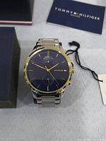 Front view of Tommy Hilfiger watch showing blue dial, gold index markers, and two tone stainless steel band with original tag.