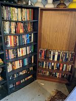 Two corner bookcases filled with a variety of hardcover books. The left side is a black painted wooden corner bookcase with shelves fully occupied by books. The right side is a wood finish corner bookcase with fewer books mostly on the lower shelves.