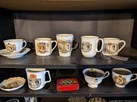 Two shelves displaying a variety of white ceramic commemorative mugs and cups with British royal family portraits and heraldic designs, along with one small red collectible tin.