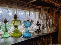 Seven vintage glass oil lamps displayed on a wooden shelf with a window background showing different colors and shapes.