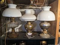 Wide view of three electrified kerosene lamps on shelf showing different glass shades and metal bases
