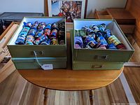 Four boxes containing assorted rolled neckties in various themes, displayed on a round wooden table, showing overall quantity and storage.