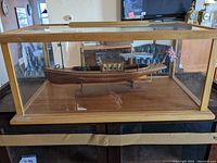 Side view of the handmade steam powered boat inside a glass display case with wooden frame and base, highlighting the polished wooden hull and small cabin area.