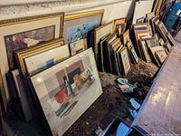 Multiple framed artworks leaning against basement wall on dirt floor