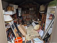 Shed interior with wooden shelves of vinyl records, assorted lamps, lamp shades, rolled textiles, ladder and storage boxes