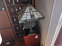 Tiffany-style stained glass table lamp with geometric patterns on shade and metal base placed on wooden furniture near window.