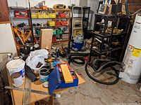 Wide view of the workshop corner showing workbench with tools, shelving with storage bins and power tools, and shop vacuum cleaner beside water heater.