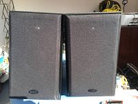 Front view of two Definitive Technology speakers with black fabric grilles and brand logo on bottom center of each grille.