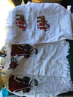 Three white Christmas dinner napkins with colorful embroidered Santa Claus figures, fringed edges visible, laid folded.