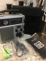 Portable silver Infinity FR-300 radio with tuning dial, speaker, and some accessories in front including batteries and cables, next to black carrying case and instruction manual.