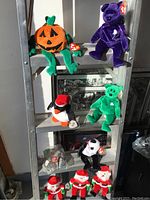 Wide view of all plush toys displayed on shelf including pumpkin, purple bear, penguin, green bear, black and white dog, three Santa Claus dolls.