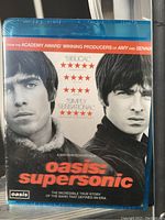 Front cover of the Oasis: Supersonic Blu-Ray showing black and white image of workers, red and orange text. Blue plastic case typical of Blu-Rays.