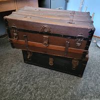 View of two stacked trunks: the top trunk has wood panels and metal corners, the bottom trunk is black with metal hardware.