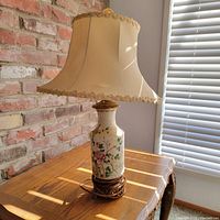 Front view of Asian ceramic vase lamp on wooden table near window with blinds, showing fabric lamp shade, painted floral blossoms on vase body, and metal stand base.