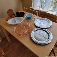 Assorted serving pieces on table including Pyrex dish, wooden box, glass cup with stand, pink glass plate, and metal trays.