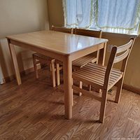Photo showing light wood rectangular breakfast table with three slatted chairs around it against a hardwood floor and near a curtained window.