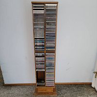 Tall wooden CD stand loaded with CDs in jewel cases, organized across multiple shelves, against a plain wall.