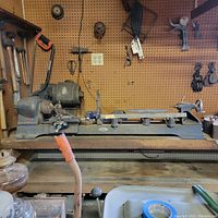 Full view of vintage Craftsman metal lathe mounted on a wooden bench with motor and toolpost, surrounded by workshop hand tools hanging on pegboard