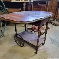 Front angle showing drop leaf tabletop with scalloped edges, turned legs, lower shelf, and both large iron wheels and small caster wheels for mobility