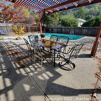 Outdoor rectangular patio table with glass top and six matching metal chairs, two swivel chairs included, placed under pergola next to swimming pool.