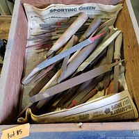 Top view of a wooden box containing several assorted metal files with wooden and plastic handles along with a measuring device, resting on old newspaper.
