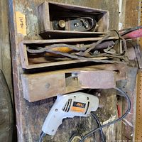 Top-down photo of woodworking tools including two wooden hand planes, four twisted steel auger bits in a wooden box, and a Skil corded drill, all placed on a worn workbench.